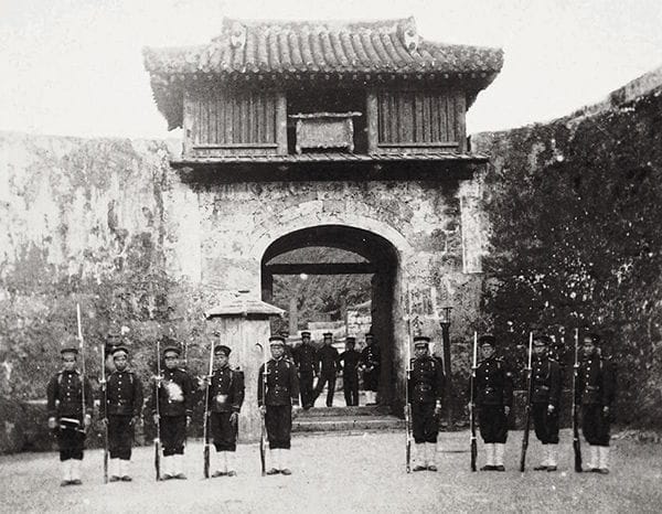 A black-and-white photograph of the Kankaimon gate of Shuri Castle, Okinawa, around 1879, with Meiji-government soldiers standing before it.