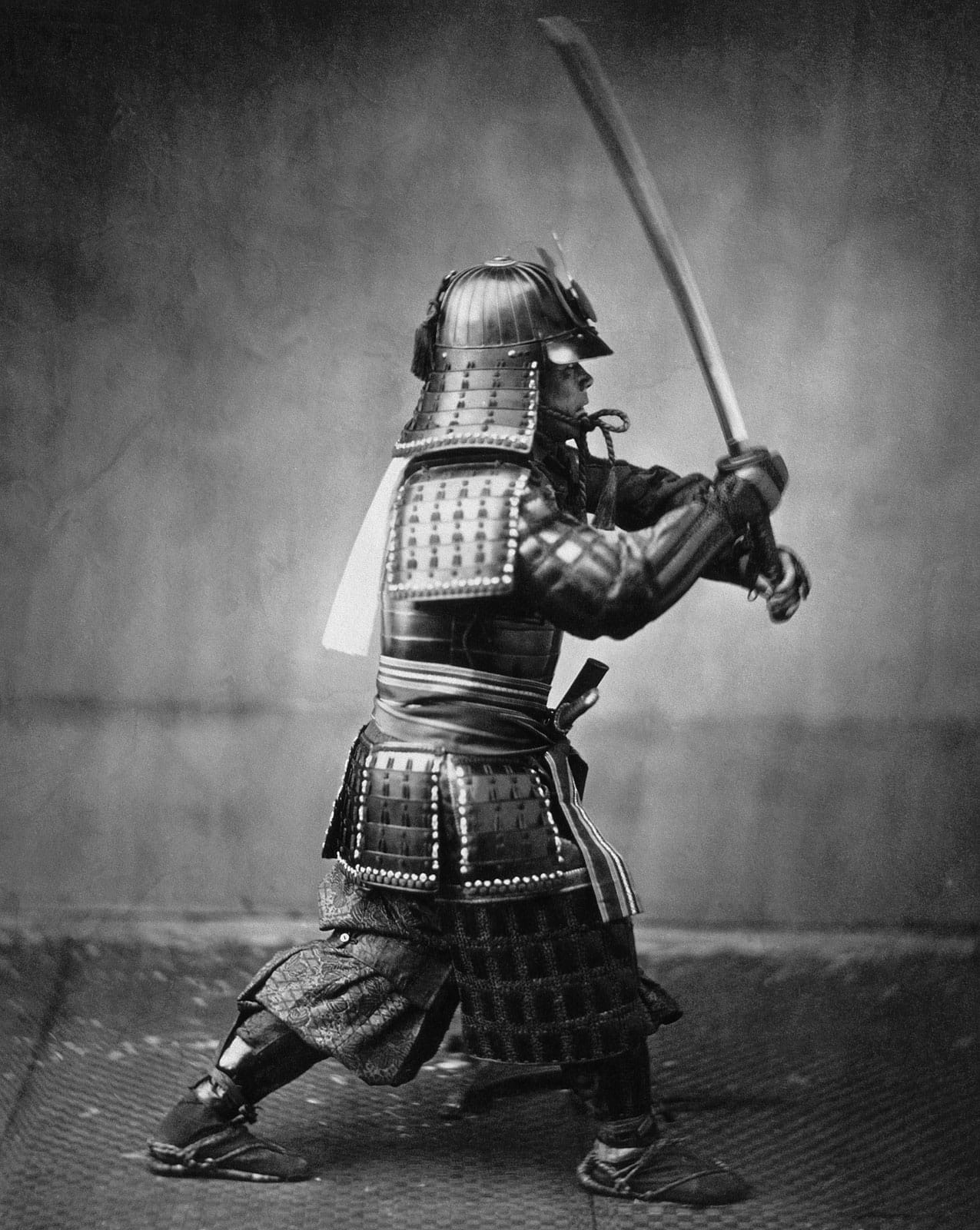 An 1860s photograph of an armoured samurai in a sword stance with the blade raised.