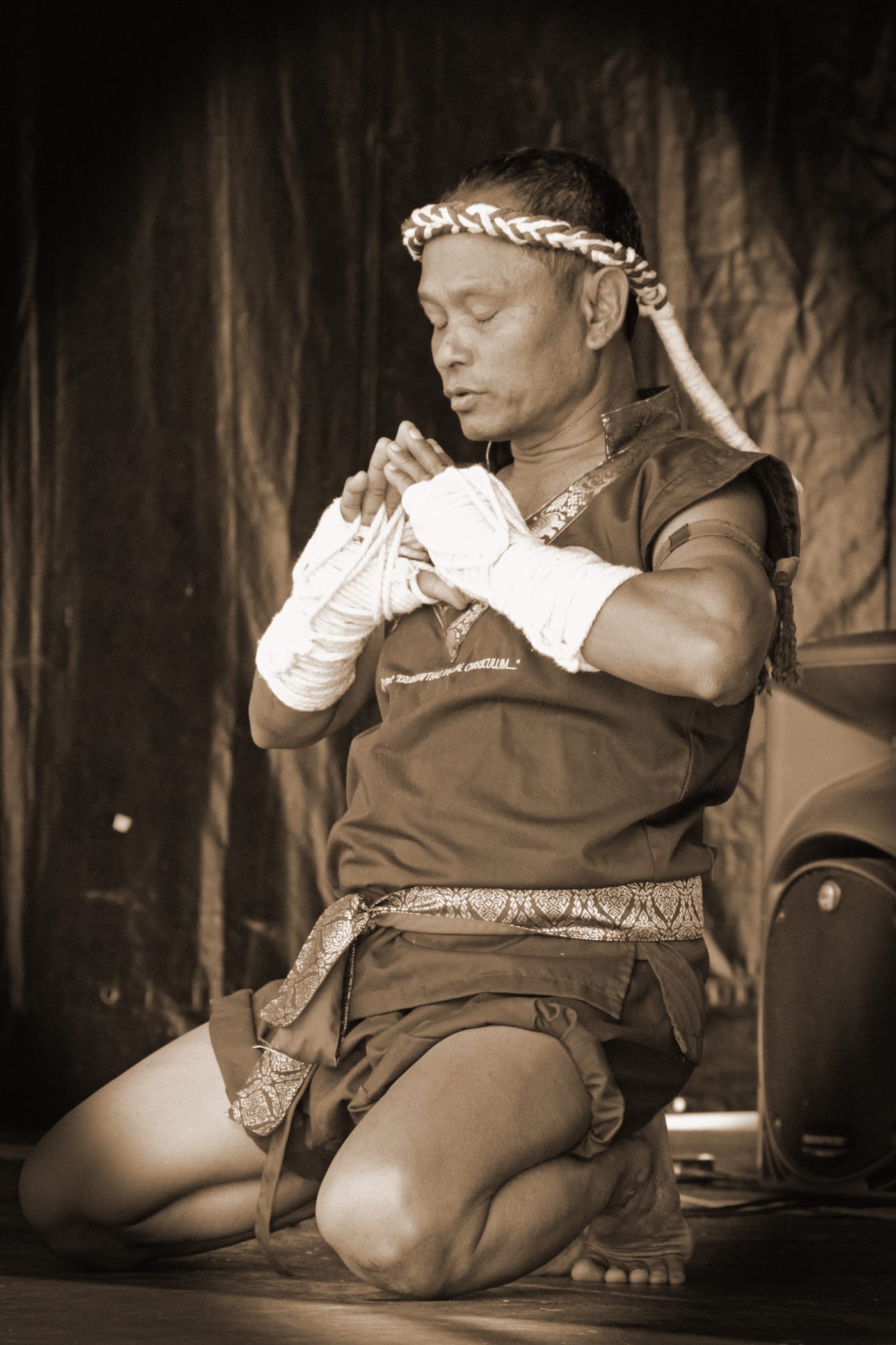 A sepia-toned photograph of a Muay Thai practitioner wrapping his hands and performing the wai khru ritual.